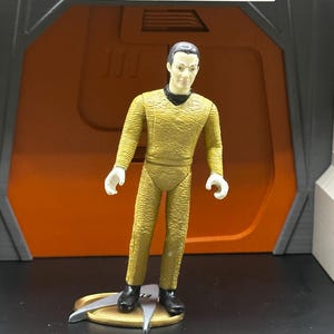 May include: A yellow Star Trek action figure of a character with short brown hair and a serious expression. The figure is standing on a silver and gold base with a Starfleet insignia.
