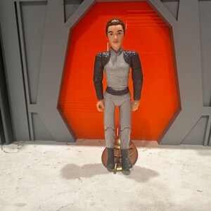May include: A grey and brown action figure of a person wearing a grey jumpsuit with a brown jacket. The figure is standing on a gold stand.