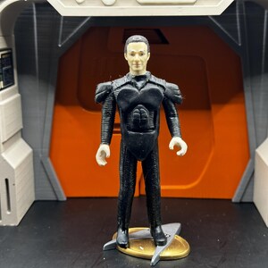 May include: A black action figure of a man wearing a black suit with a textured chest piece. The figure is standing on a gold stand with a silver star-shaped base.