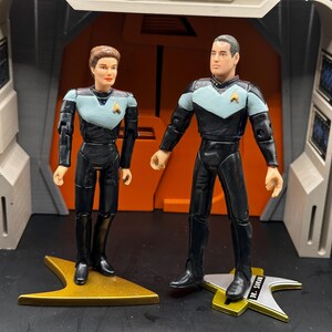 May include: Two action figures of Star Trek characters, a woman with short brown hair wearing a blue and black uniform and a man with short brown hair wearing a black and blue uniform, standing on gold and silver star-shaped bases.