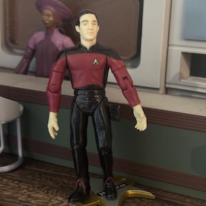 May include: A Star Trek action figure of the character Data, featuring a pale complexion, dark hair, and a maroon and black uniform with a gold Starfleet insignia on the chest. The figure stands on a gold-colored base with the name Data printed on it.