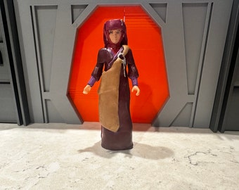 May include: A burgundy and tan action figure of a female character wearing a hooded robe and a long, flowing dress. The figure is standing in front of a red and gray background.