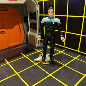 May include: A black and blue Star Trek action figure of a character wearing a black uniform with a blue collar and a silver badge. The figure is standing on a gold and silver Starfleet insignia.