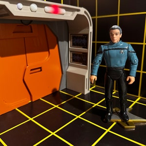May include: A blue and black action figure of a Star Trek character standing on a gold-coloured base. The figure is in front of a model of a spaceship door.