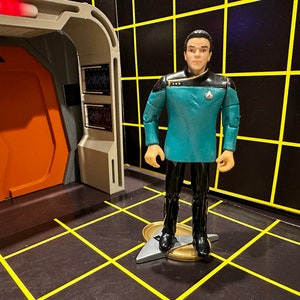 May include: A teal blue Star Trek uniform with black trim and a gold Starfleet insignia. The figure is standing on a silver and gold Starfleet insignia base.