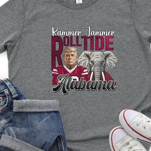 以下が含まれることがあります： グレーのTシャツに、赤と白のフットボールジャージを着た男性のグラフィックがプリントされています。ジャージには「Roll Tide」と「Alabama」の文字が書かれています。男性は腕を組み、真剣な表情をしています。男性の右にはゾウがいます。グラフィックの上には「Rammer Jammer」の文字が書かれています。