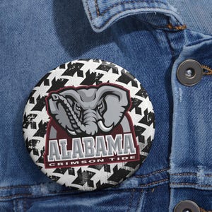 May include: A round button featuring the Alabama Crimson Tide logo. The button has a houndstooth pattern in black and white. The logo includes an elephant graphic and the words "ALABAMA CRIMSON TIDE" in red and white.