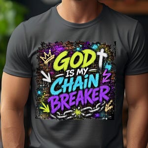 May include: A charcoal gray crew neck t-shirt featuring a vibrant graffiti-style graphic on the chest. The design displays the text "GOD IS MY CHAIN BREAKER" in bright yellow, blue, and purple, accompanied by a crown, a cross, and a broken chain.