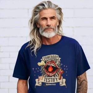 May include: Navy blue t-shirt with a graphic design featuring open lips with a flame, a cross, and the words "SOMEBODY TESTIFY Matthew 10:33". The design is in shades of red, white, and gold.