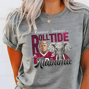 以下が含まれることがあります： 赤、白、青のグラフィックがプリントされたグレーのTシャツ。赤い白のフットボールジャージを着た男性、ゾウ、そして「Rammer Jammer Roll Tide Alabama」の文字が描かれている。
