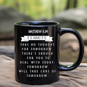 May include: Black ceramic mug with a curved handle. The mug features the text "MATTHEW 6:34" and the phrase "IT IS WHAT IT IS" above a longer quote about not worrying about tomorrow. The mug is sitting on a wooden surface.
