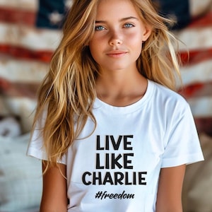 May include: White t-shirt with the text "LIVE LIKE CHARLIE" in a bold, black font, with the hashtag "#freedom" below. The shirt is worn by a person with long blonde hair and blue eyes.