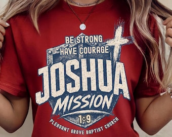 May include: A dark red short-sleeve t-shirt featuring a graphic design with a shield and cross. The text reads BE STRONG HAVE COURAGE JOSHUA MISSION 1:9 PLEASANT GROVE BAPTIST CHURCH in a distressed, off-white and dark blue color scheme.