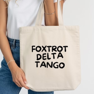 May include: Cream-colored tote bag with the words "FOXTROT DELTA TANGO" printed in bold black letters. The bag is held by a person wearing a white t-shirt and blue jeans.