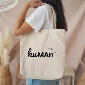 May include: Beige canvas tote bag with black text that reads "human" with "man" in a bold font. The words "THAT'S IT" are in a semi-circle around the right side of the word "human". The bag has two handles.