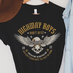 Puede incluir: Camiseta negra con un diseño gráfico de un águila con una calavera en sus garras. El texto "Highway Boys Don't Rest" está por encima del águila y "American Heartbreak And Don't Hang Hats 'Til They're Home" está por debajo del águila. Los números 20 y 22 están a ambos lados del águila.