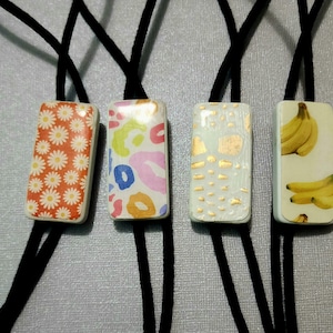 May include: Four rectangular pendants with black cords. The pendants feature various designs: a daisy pattern, colorful abstract shapes, gold leaf accents, and a bunch of bananas. The pendants are likely for necklaces or similar accessories.
