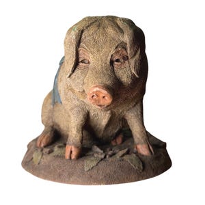 May include: A brown, textured pig figurine in a seated position, featuring a pinkish snout, drooping ears, and a small blue accent on its back. The pig is positioned on a circular, earth-toned base that includes sculpted leaf details on the ground.