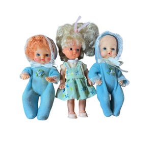May include: Three dolls standing together. Two dolls wear light blue onesies with white lace bonnets and small floral accents. The center doll has blonde hair, wears a light green dress with a white shirt, and has pink shoes.