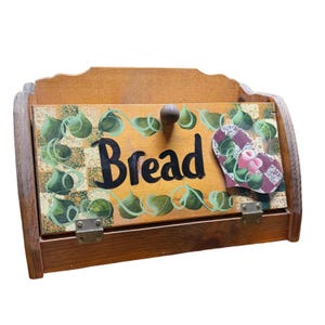 Puede incluir: Una panera de madera con una puerta frontal abatible que muestra la palabra Bread en letras negras. La puerta está decorada con un patrón pintado de hojas verdes sobre un fondo a cuadros, con un pequeño pomo de madera y bisagras metálicas.