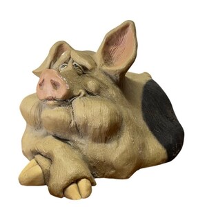May include: A tan-colored pig figurine with a pink snout, large ears, and a prominent black patch on its side. The pig is depicted in a resting position with its front hooves crossed, featuring detailed facial expressions and textured skin.