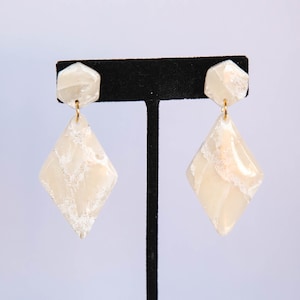 May include: A pair of cream-colored, geometric earrings. Each earring features a hexagonal top and a diamond-shaped pendant. The earrings have a marbled effect with gold-colored hooks, displayed on a black stand against a white background.