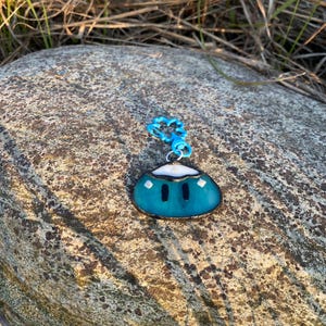 Puede incluir: Un dije turquesa con una parte superior blanca y dos formas negras parecidas a ojos. Está unido a una cadena azul. El dije está sobre una roca gris, con hierba seca en el fondo.