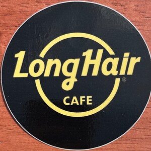 May include: A circular black sticker featuring the words "LongHair CAFE" in yellow. The text is within a yellow circle, with "LongHair" above "CAFE". The sticker has a white border.
