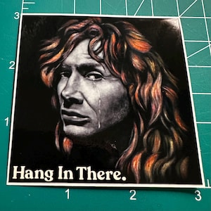 May include: Square sticker featuring a grayscale portrait of a person with long, wavy, reddish-brown hair and a tear rolling down their cheek. The text "Hang In There." is printed in white at the bottom of the sticker.