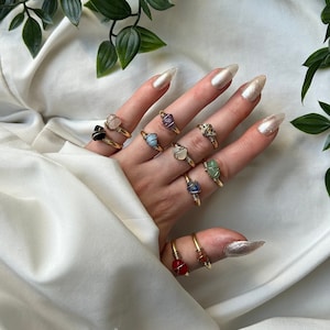 Könnte beinhalten: Eine Hand trägt eine Vielzahl von goldenen Ringen mit verschiedenen farbigen Edelsteinen. Die Ringe sind mit kleinen, unregelmäßig geformten Steinen besetzt. Die Hand ruht auf einem weißen Stoffhintergrund mit grünen, blättrigen Ranken.
