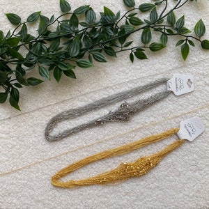 May include: Two silver and gold chain necklaces on a white surface. The necklaces are layered and have a delicate, feminine look. The silver necklace has a tag that says "Flauschstern" and the gold necklace has a tag that says "Flauschstern".