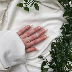 Könnte beinhalten: Eine Hand mit weißem Nagellack und kleinen Perlenakzenten auf den Nägeln trägt zwei Ringe. Ein Ring ist ein dünnes Band mit einer einzelnen Perle, der andere ist ein dickeres Band mit mehreren Perlen. Die Hand ruht auf einem weißen Stoffhintergrund mit grünen, blättrigen Ranken.