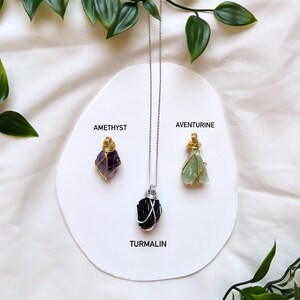 May include: Three necklaces with different colored crystals, amethyst, aventurine, and tourmaline, are displayed on a white circular surface with the names of the crystals written below each one. The necklaces are silver chains with gold wire wrapped around the crystals.