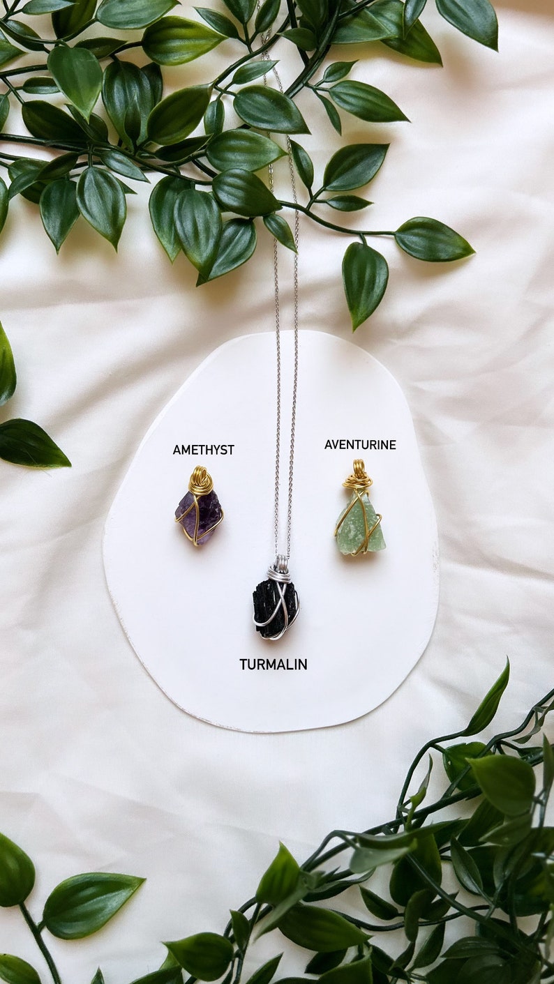 May include: Three necklaces with different colored crystals, amethyst, aventurine, and tourmaline, are displayed on a white circular surface with the names of the crystals written below each one. The necklaces are silver chains with gold wire wrapped around the crystals.