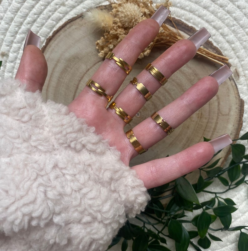 May include: A hand wearing a set of six gold-toned rings. The rings are all different styles and sizes, and they are stacked on each other on the fingers.