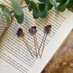 Könnte beinhalten: Fünf goldene Haarnadeln mit Amethyst-Steinen. Die Amethyst-Steine sind lila und die Nadeln sind golden. Die Nadeln liegen auf einem Buch mit Text über Energie und Schwingungen.