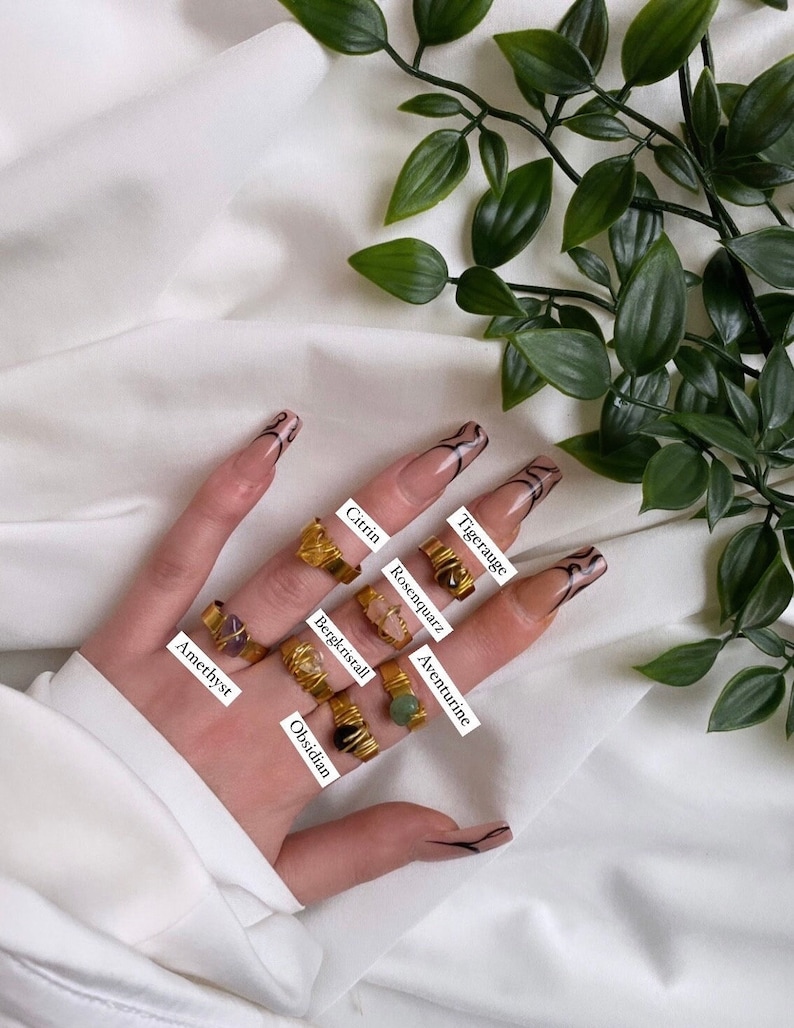 May include: A hand wearing seven gold rings with different colored gemstones. The stones are labeled with their names: Amethyst, Obsidian, Bergkristall, Citrin, Tigerauge, Rosenquarz, and Aventurine.