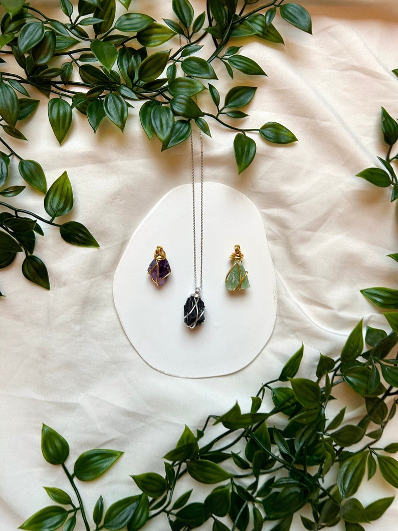 May include: Three wire-wrapped crystal necklaces on a white surface. The necklaces feature a purple amethyst, a black crystal, and a green fluorite.