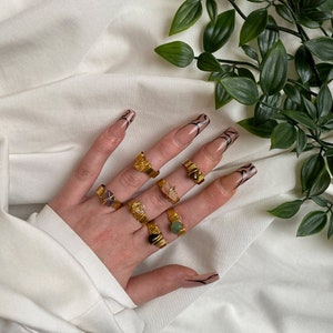 May include: A hand wearing multiple gold rings with different colored gemstones. The rings are made of wire wrapped around the stones. The hand is on a white fabric background with green leaves.