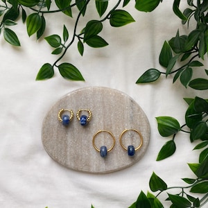 May include: A set of gold hoop earrings with blue gemstone accents. The earrings are displayed on a marble surface with green foliage in the background.