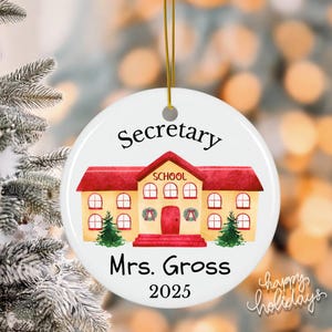 May include: A white, round Christmas ornament with a schoolhouse design. The ornament features the word "Secretary" at the top, the name "Mrs. Gross" below the school, and the year "2025" at the bottom. The words "happy holidays" are written in cursive.