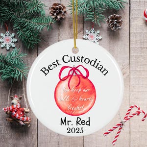 Può includere: Ornamento natalizio in ceramica bianca con la scritta "Best Custodian" e "Mr. Red 2025". L'ornamento presenta una palla di Natale rossa in stile acquerello con un fiocco rosso. Il testo "You keep our halls & hearts bright!" è scritto sulla palla.