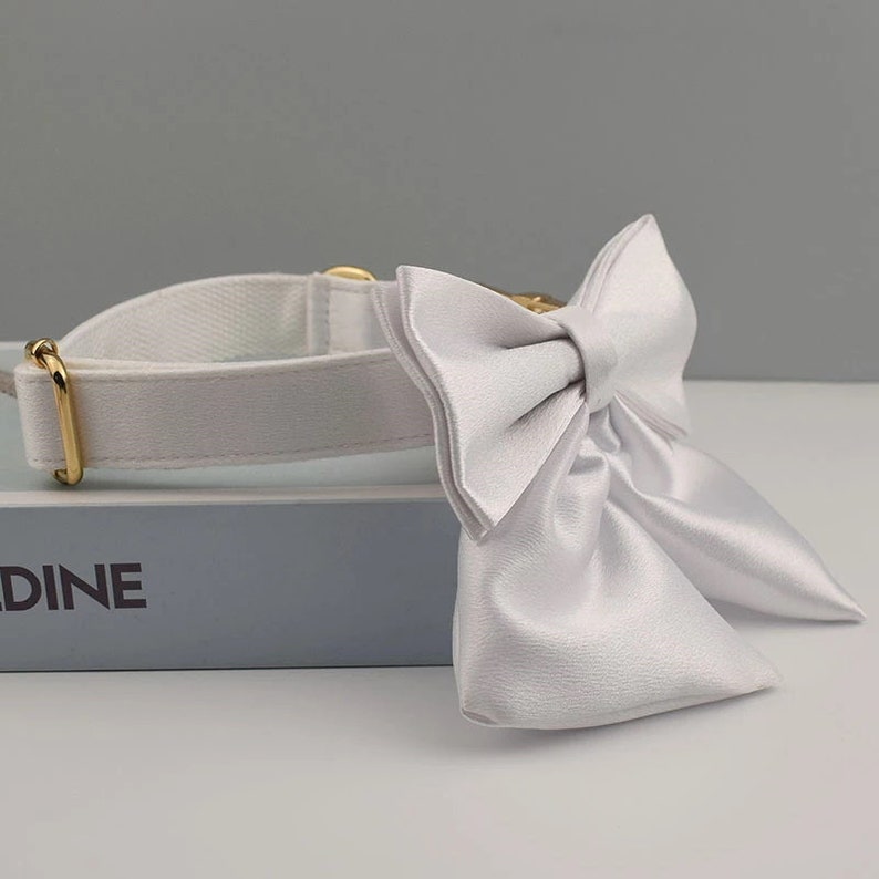May include: White dog collar with a large white satin bow. The collar is made of a white canvas material and has a gold metal buckle.