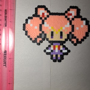May include: Pixel art figure of a character with pink hair, a purple dress, and black accents. The figure is made of small, colorful beads. A pink ruler with inch measurements is on the left side of the image.