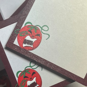 May include: Two red tomato-shaped sticky notes with a brown border. Each tomato has a green stem and a white face with a wide open mouth and sharp teeth. The tomatoes have a cartoonish angry expression.