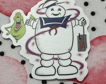 May include: A white sticker featuring the Stay Puft Marshmallow Man in a sailor outfit, holding a detonator. A green cartoon ghost is to the left. Purple swirls and starbursts surround the characters.