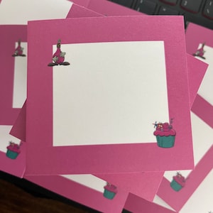May include: A stack of pink note cards with a white interior. Each card features a whimsical illustration of a cupcake with pink frosting and a bottle with a red cap.