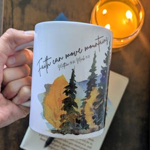 May include: White ceramic mug with the text "Faith can move mountains" and a watercolor landscape of trees and a mountain. The mug is held by a person's hand. A lit candle and a book are in the background.