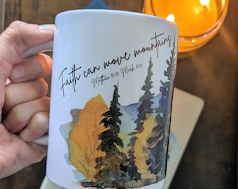 May include: White ceramic mug with the text "Faith can move mountains" and a watercolor landscape of trees and a mountain. The mug is held by a person's hand. A lit candle and a book are in the background.