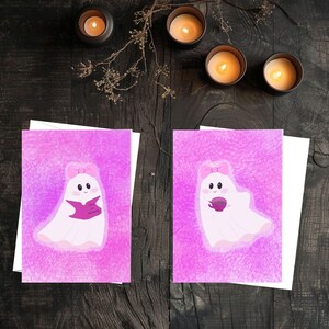 May include: Two greeting cards with a pink ghost illustration. The ghosts have a pink bow and are holding a book and a cup. The background is a textured pink. Lit candles and dried sprigs are in the background.